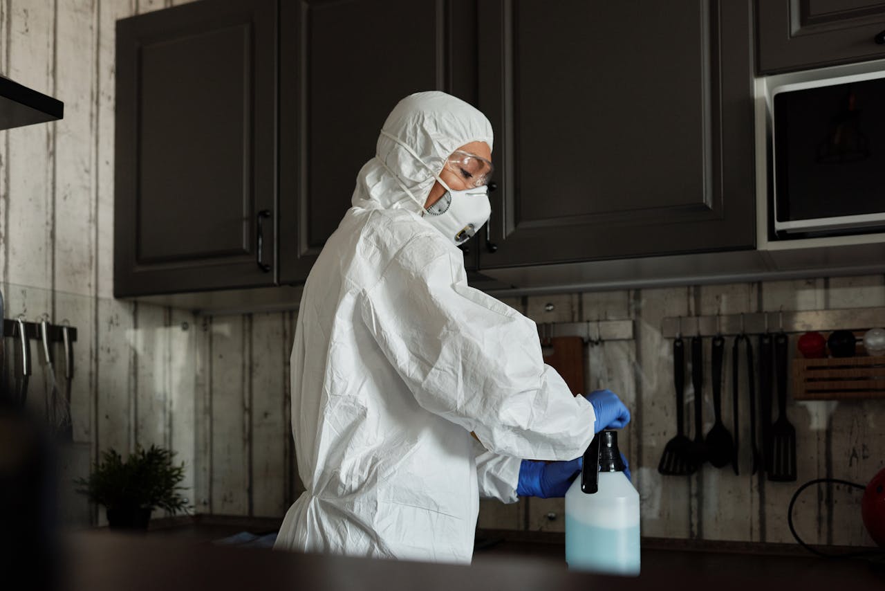 services-01 Healthcare worker in PPE uses disinfectant spray in a kitchen setting during COVID-19 pandemic.