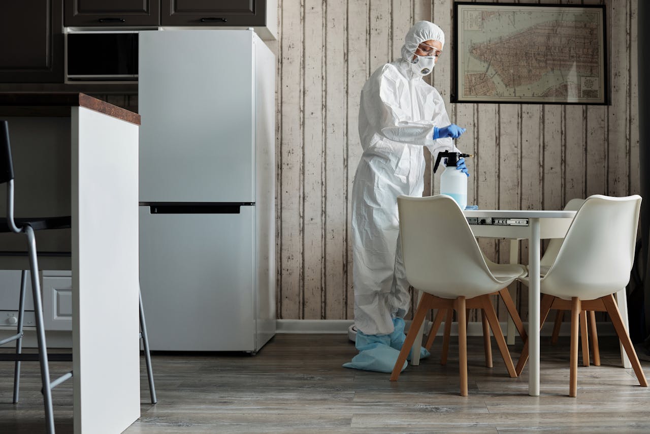 services-02 Person in hazmat suit disinfecting a modern kitchen table with spray bottle for hygiene.